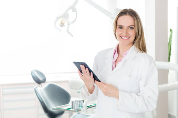 Horizontal color image of female dentist holding digital tablet in dental clinic office, smiling and looking at camera. Female doctor wearing white uniform. Dental equipment in the background.