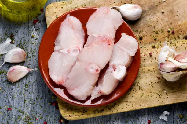 high-angle shot of some slices of raw monkfish on a wooden shopping board, some garlics and a cruet of olive oil on a rustic wooden table
