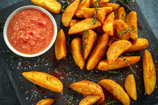Fried potato wedges with hot salsa sauce, herbs on stone board.