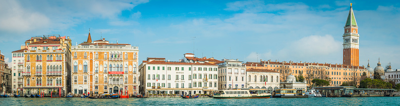Panoramic view across the busy waterway of the Grand Canal with its crowded water buses and taxis plying their trade between the gondolas and working boats of Venice, overlooked by the iconic Campanile bell tower in St. Mark’s Square and the colourful stucco villas and hotels of this iconic Italian city.