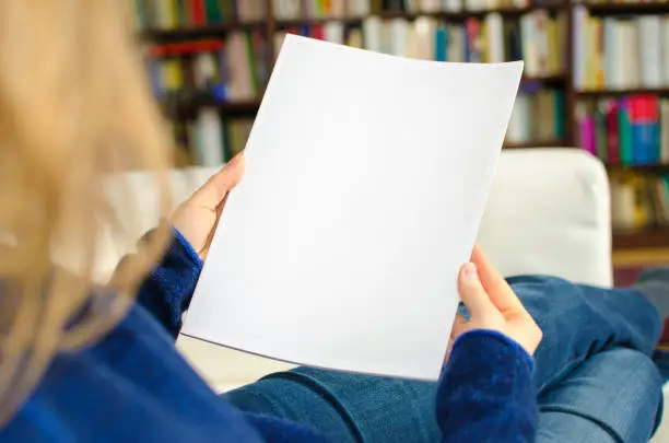 Blond woman holding a blank brochure or book cover with copy space. The photo was taken indoors in a domestic room by photographing over the woman's shoulder.