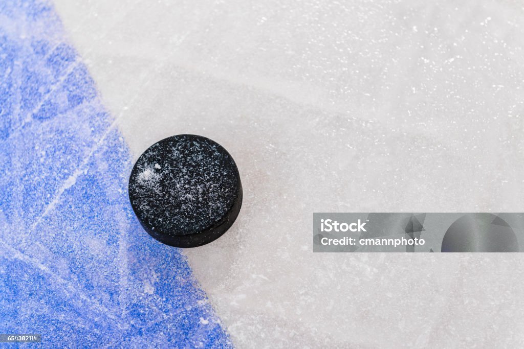 Ice Hockey puck next to Blue Line in rink Looking down on a ice hockey puck sitting next to the Blue Line with marred up ice from the skate blades. The Blue Line separates the attacking and defending zones from the neutral zone in the center of the rink. Hockey Stock Photo