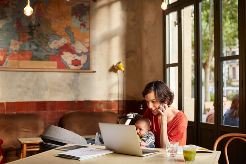 Mother carrying baby using phone while looking in laptop at table in restaurant