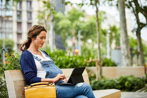 Pregnant woman using laptop while sitting on bench in park