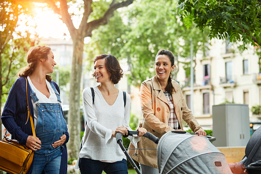 Happy pregnant woman talking to friends pushing baby strollers on sunny day