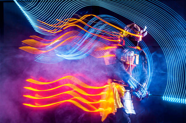 Young man dressed in a samurai costume with glowing LED light strip. The young man is performing the dance leaving traces in the form of luminous stripes. He is waving the LED staff and moving in dark. Motion blur on long exposure. Studio shooting with smoke on black background with a rising red sun at white in a middle of composition