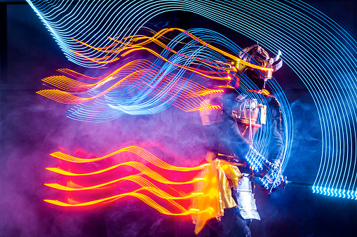 Young man dressed in a samurai costume with glowing LED light strip. The young man is performing the dance leaving traces in the form of luminous stripes. He is waving the LED staff and moving in dark. Motion blur on long exposure. Studio shooting with smoke on black background with a rising red sun at white in a middle of composition