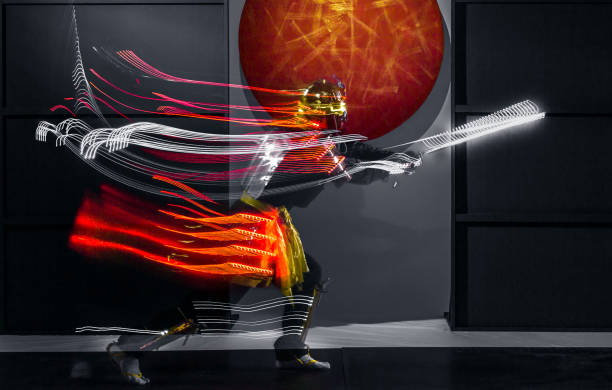 Young man dressed in a samurai costume with glowing LED light strip. The young man is performing the dance leaving traces in the form of luminous stripes. He is waving the LED staff and moving in dark. Motion blur on long exposure. Studio shooting with smoke on black background with a rising red sun at white in a middle of composition