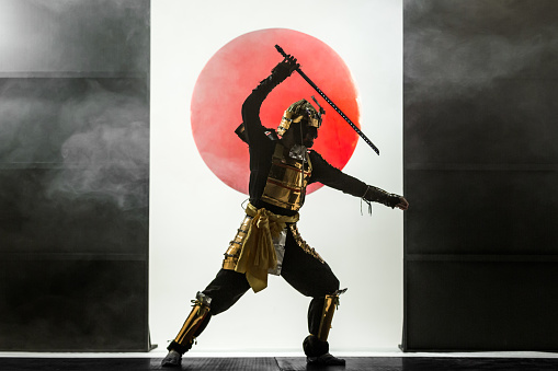 Young man dressed in samurai costume. The young man is posing waving sword. Studio shooting with smoke on black background with a rising red sun at white in a middle of composition