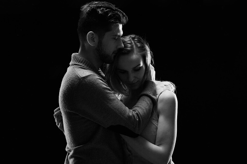 Young serious couple hugging and standing against black background