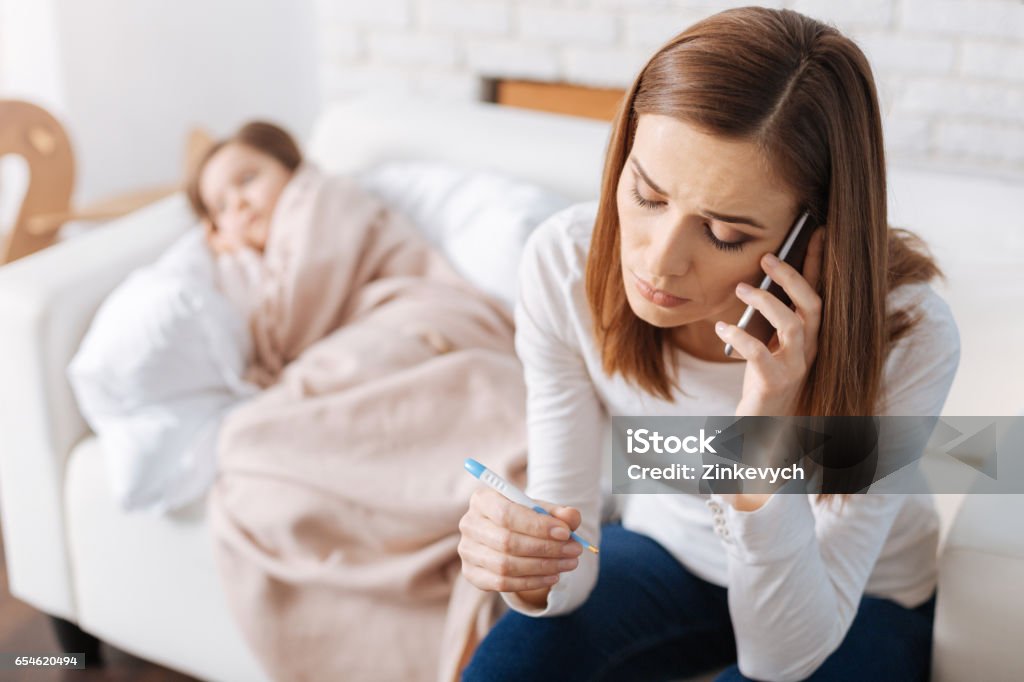 Cheerless mother calling a doctor Urgent help. Cheerless troubled mother holding thermometer and calling a doctor while her ill daughter sleeping in the background Doctor Stock Photo