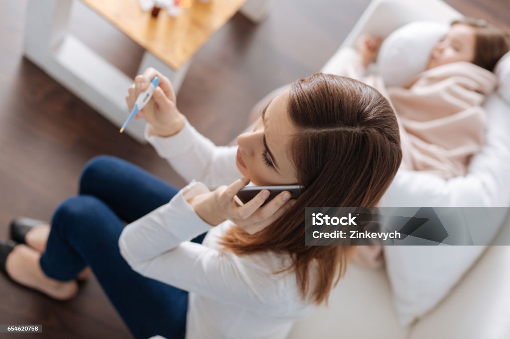 Cheerless caring mother calling a doctor High temperature. Caring cheerless moter calling a doctor and holding thermometer while her sick little daughter lying in the background Child Stock Photo