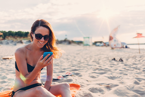 Smiling woman sunbathing at the beach and using smartphone