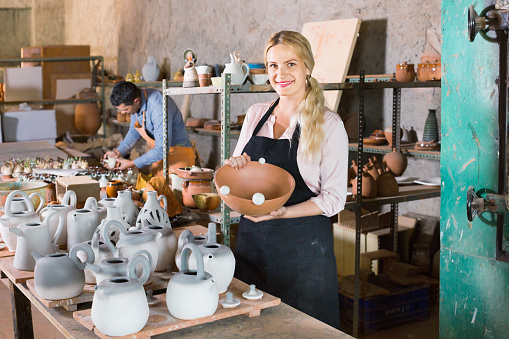 Portrait of cheerful efficient woman pottery worker with ceramic crockery in hands in studio