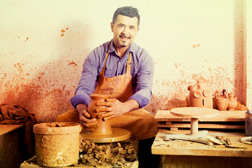 happy european artisan man creating ceramic piece on spinning pottery wheel in workshop