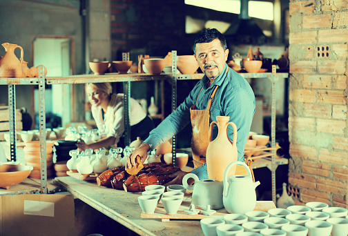 two cheerful artisans having ceramics in hands and standing in workshop