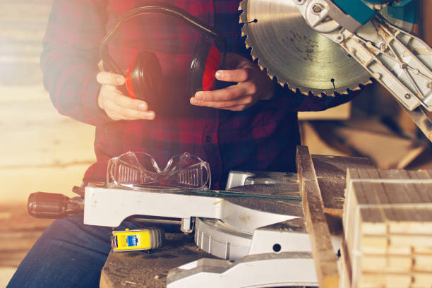 istock Sawmill workplace concept. Workman dressed in the checkered shirt holding protective headphones near circular saw. 654826312
