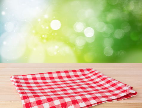 Red picnic cloth folded on wooded table with green nature blurred background.Empty napking advertisement concept.