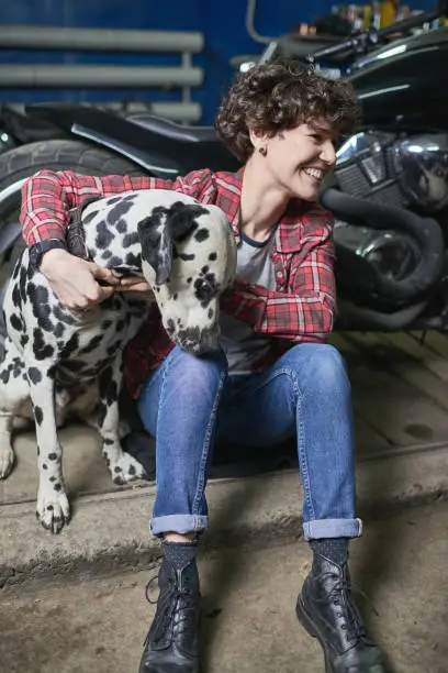 Optimistic young woman in masculine clothes sitting in garage and embracing her dog