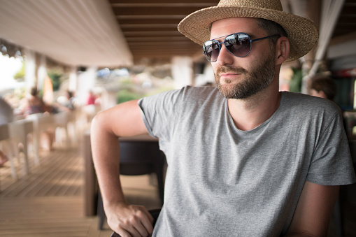 Close-up shot of a handsome young man with sunglasses and a hat sitting at the beach bar and enjoying the tropical view.