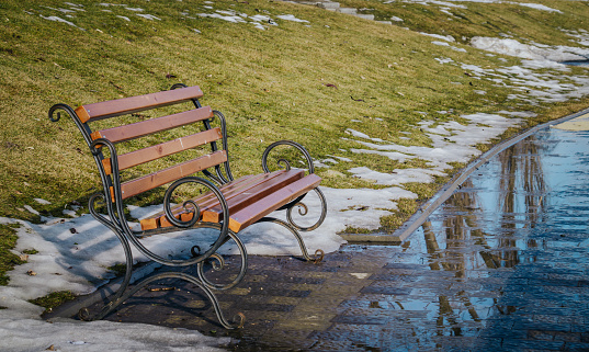 Spring urban landscape. Garden bench in a deserted city Park. Melted snow and puddles