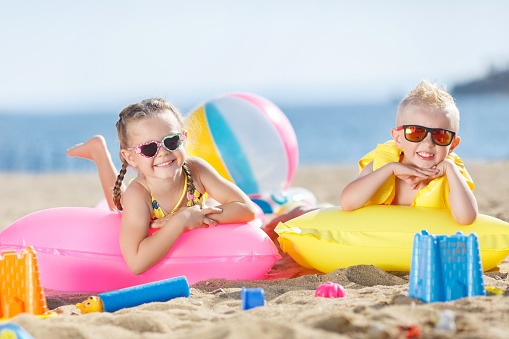 Little girl with pigtails and a little blond boy with short hair,wearing dark sun glasses, spend time together on the sandy beach,lie side by side on air mattresses for swimming on the blue ocean,mattresses yellow and pink