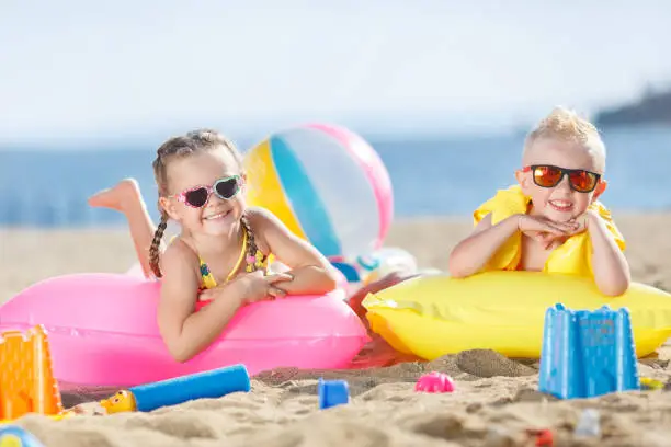 Little girl with pigtails and a little blond boy with short hair,wearing dark sun glasses, spend time together on the sandy beach,lie side by side on air mattresses for swimming on the blue ocean,mattresses yellow and pink