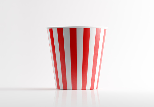 Red and white striped popcorn bucket. Popcorn bucket is empty and made of paper. Vertically stripped. Lit from the right upper corner of composition and casting shadows on a white reflective surface. Horizontal composition with copy space.