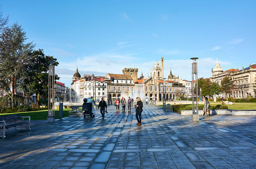 People enjoy the sunshine after rain. the square is still wet. Big central square in Braga with several fountains. Sunny day in spring with blue sky and small white clouds. Backlit scenery with lens flare