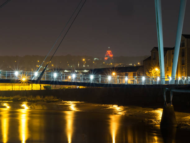 Lancaster City at Dawn with Millenium bridge over River Lune and Ashton Memorial lit up with red lights.  Orange street lights reflect in the river and white lights on the bridge sparkle.