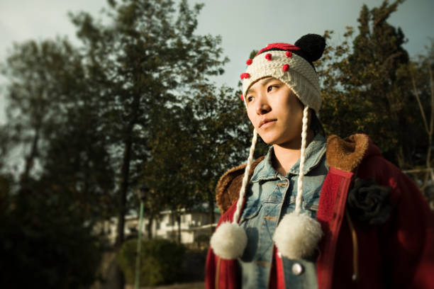 Outdoor image of an Asian girl standing in a park and enjoying fresh air in winter. She is in a hoodie, denim shirt and a fancy woolen cap. One person, horizontal composition with copy space and selective focus.