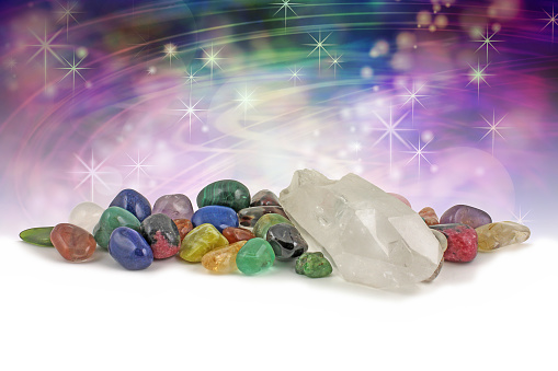 Large clear crystal terminated quartz specimen surrounded by a selection of chakra colored crystals on a magical purple and green background with copy space