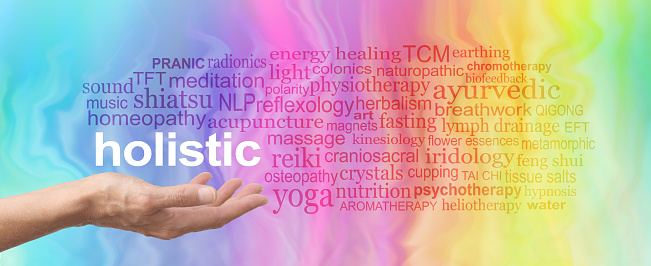Female hand held palm up the word HOLISTIC in white above surrounded by a relevant word cloud on a rainbow coloured marble effect background