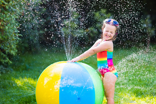 Funny laughing little girl in a colorful swimming suit playing with toy ball garden sprinkler with water splashes having fun in the backyard on a sunny hot summer vacation day