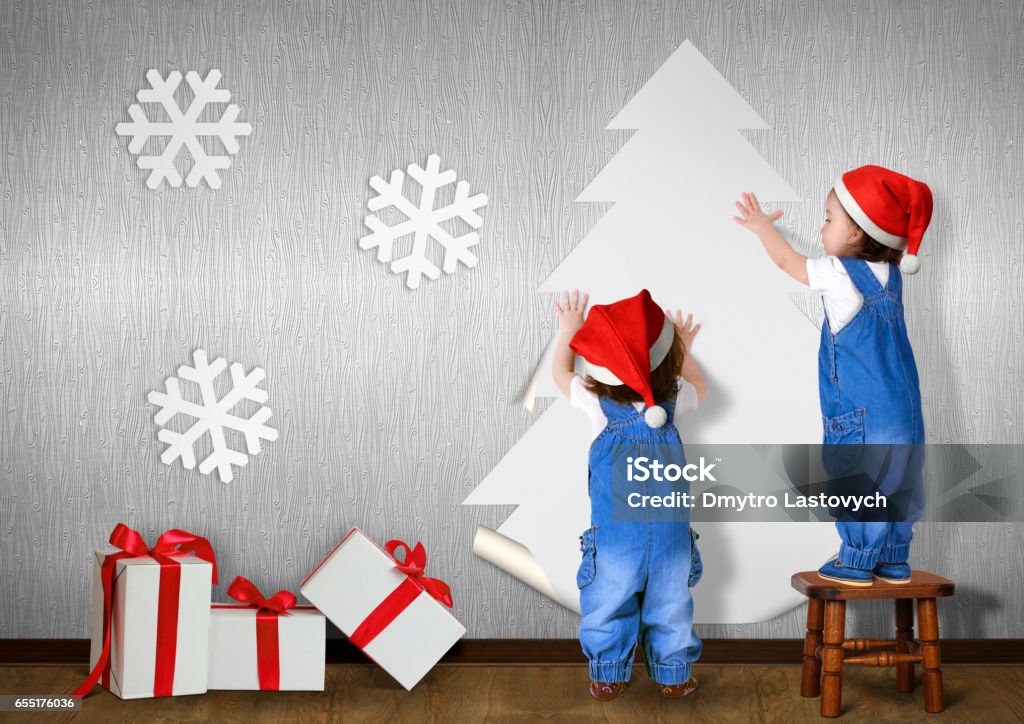 Little twins dressed Santa hat, glue Christmas tree on wall at home, xmas concept Bizarre Stock Photo