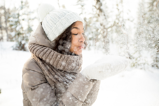 Portrait of happy teenage Latin American girl wearing white hat, scarf and jacket having fun blowing snow off her hands in winter
