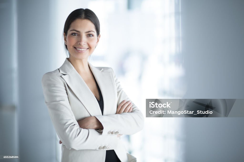 Smiling Female Business Leader With Arms Crossed Closeup portrait of smiling beautiful middle-aged business woman wearing jacket and standing in light office hall with her arms crossed Businesswoman Stock Photo