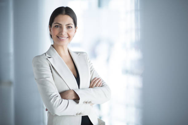 Smiling Female Business Leader With Arms Crossed Closeup portrait of smiling beautiful middle-aged business woman wearing jacket and standing in light office hall with her arms crossed saleswoman stock pictures, royalty-free photos & images