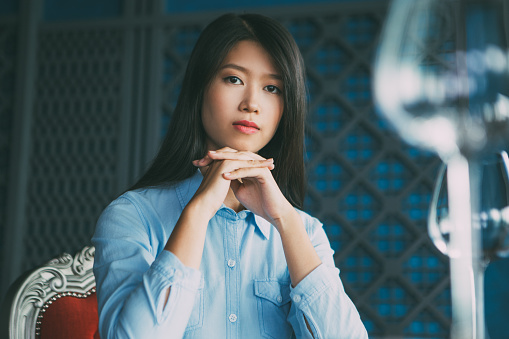 Portrait of pretty young Asian woman with her hands under chin sitting at table in luxury restaurant, looking at camera