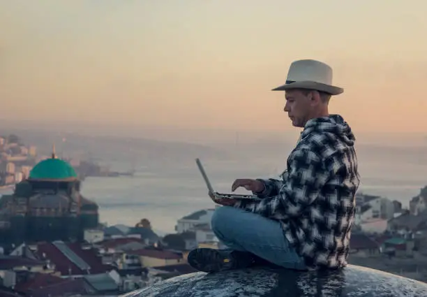 Serious Man in a hat and a plaid jacket and jeans sitting and Working with white laptop on  background overview the City at Dawn