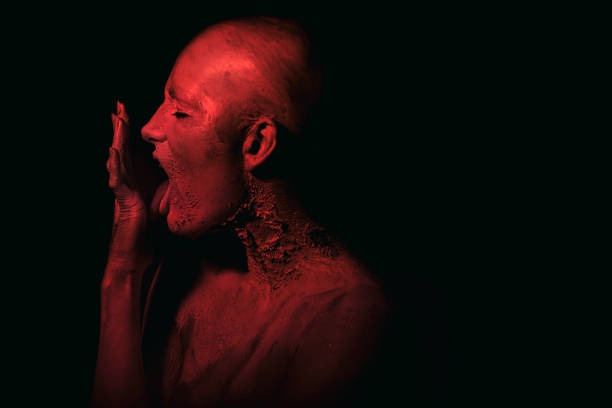 Portrait of a female bald person, her head is covered with white paint, lit with a red light. She is licking her hand.