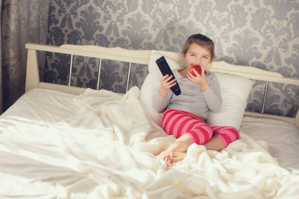 istock Little girl lying in bed with remote control TV and Apple 655352686