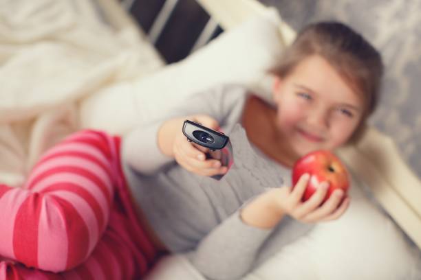 Little girl lying in bed with remote control TV and Apple