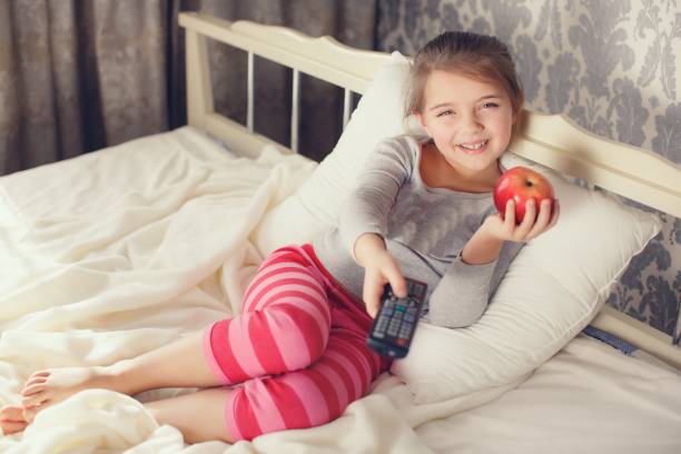 istock Little girl lying in bed with remote control TV and Apple 655352698