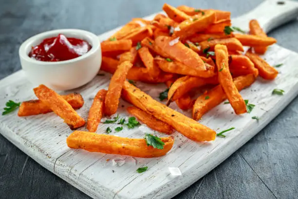 Healthy Homemade Baked Orange Sweet Potato Fries with ketchup, salt, pepper on white wooden board.