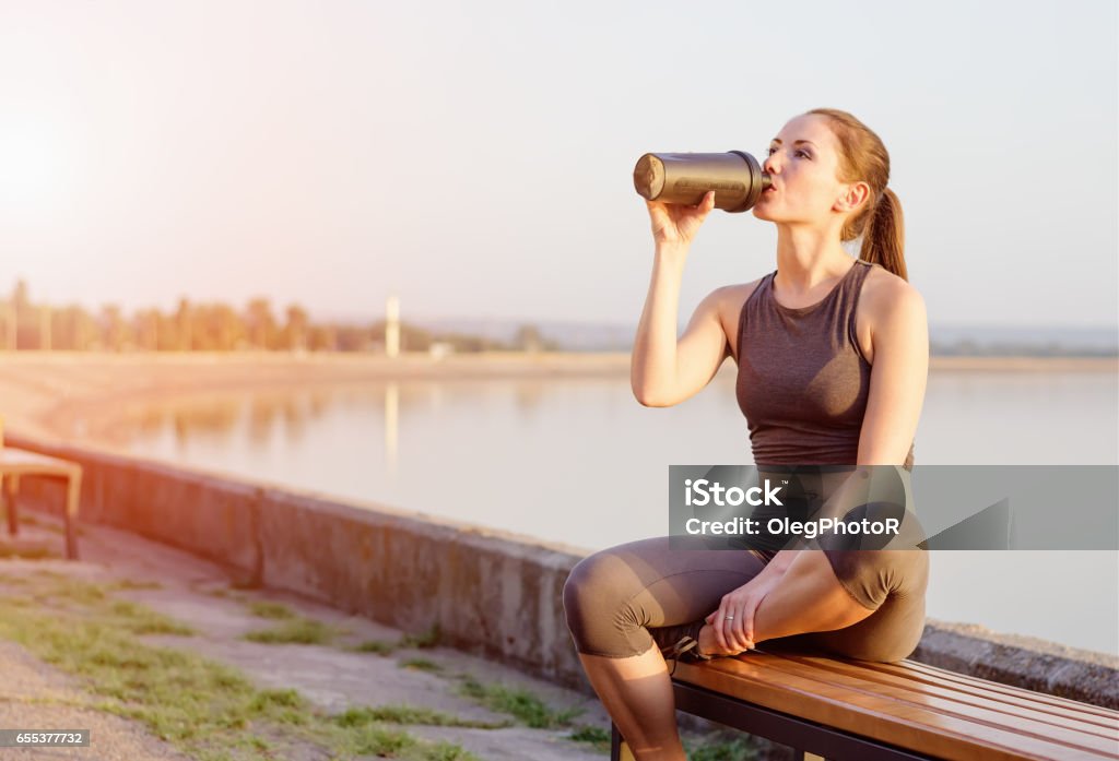 young girl drinks a protein cocktail from a schweeter after jogging young girl of Caucasian appearance drinks a protein cocktail from a shaker after jogging in the open air. The bright sun illuminates it. Protein Stock Photo