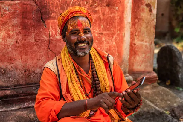 Sadhu - indian holyman using the mobile phone. In Hinduism, sadhu, or shadhu is a common term for a mystic, an ascetic, practitioner of yoga (yogi) and/or wandering monks. The sadhu is solely dedicated to achieving the fourth and final Hindu goal of life, moksha (liberation), through meditation and contemplation of Brahman. Sadhus often wear ochre-colored clothing, symbolizing renunciation.