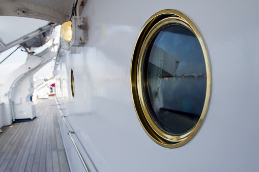 View of a round porthole with golden rim on board of a white painted luxurious yacht