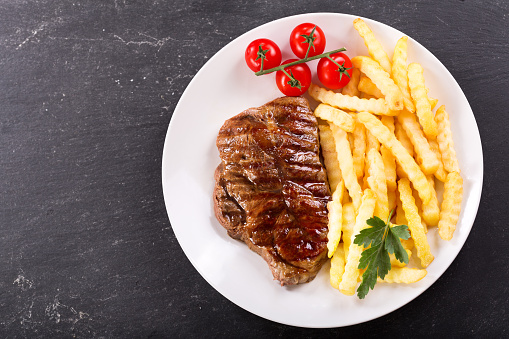 plate of grilled meat with french fries on dark background, top view