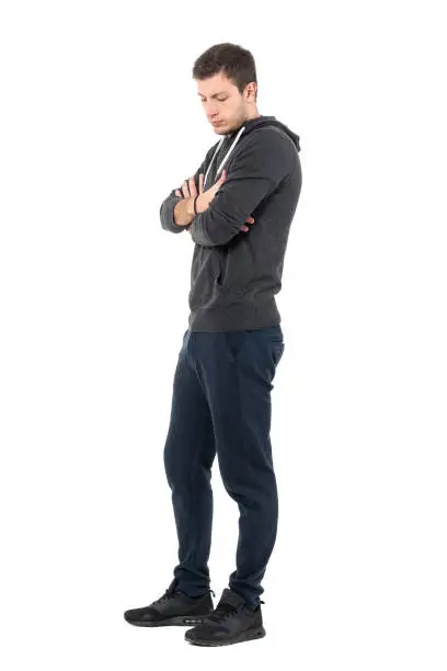 Side view of sad young man in sportive clothing looking down with crossed arms. Full body length portrait over white studio background.